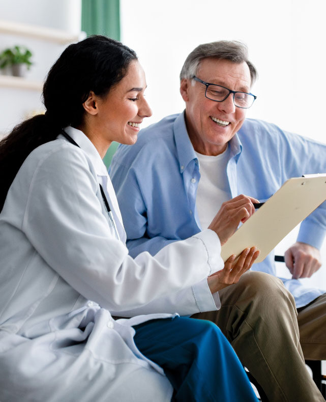 A physician in a white coat smiling with an older patient while reviewing a clipboard together