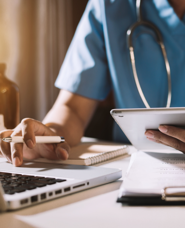 Clinician in scrubs taking notes beside a laptop while holding a tablet